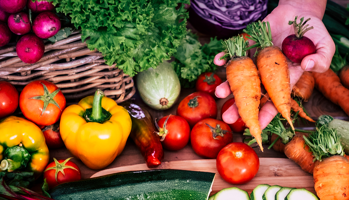 A hand holds carrots and a beet among a spread of other vegetables.