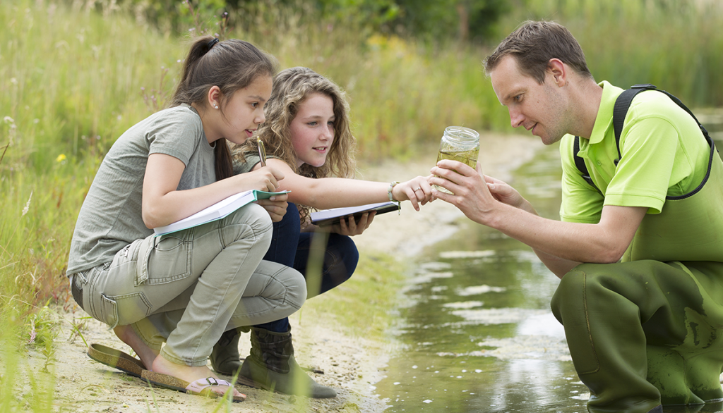 a man wearing waders shows a specimen in a jar to two young learners