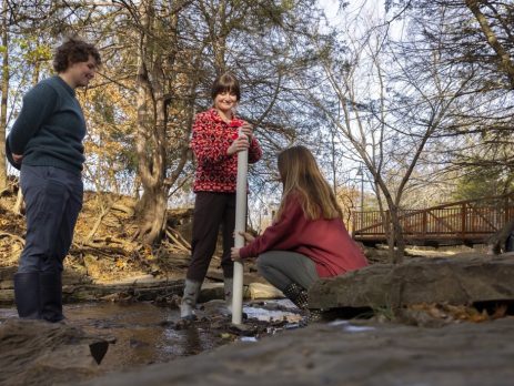 Three women stand in a shallow creekbed. One watches as the other two use a large plastic pipe to collect water samples.