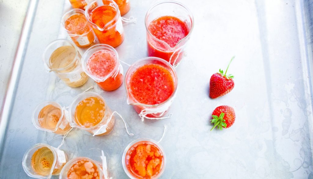 Multiple glass containers with orange and red fruit liquids in them sit on a table.