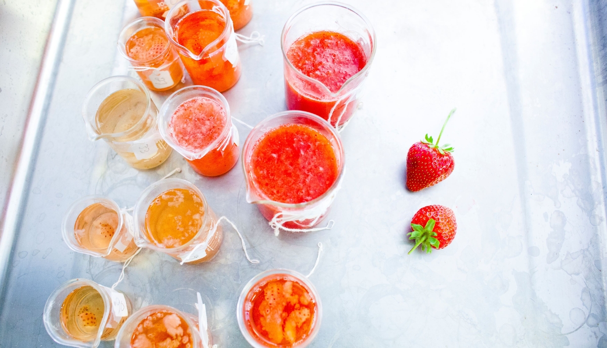 Multiple glass containers with orange and red fruit liquids in them sit on a table.