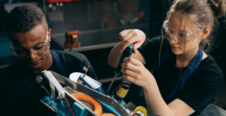two teens wearing goggles and using tools in a workshop setting