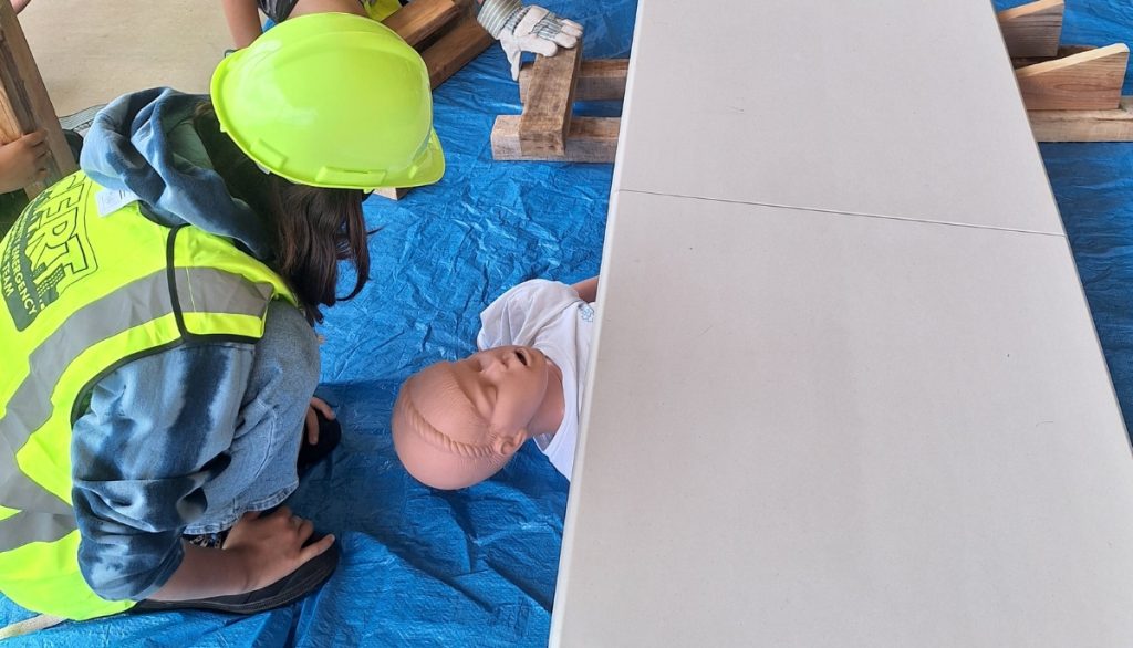 Five young people wearing neon green hard hats and safety vests participate in a hands-on training exercise. Two individuals kneel on a blue tarp, practicing emergency response techniques on a medical manikin, while others observe and take notes around a white folding table.