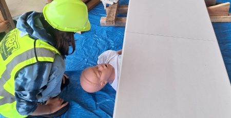 Five young people wearing neon green hard hats and safety vests participate in a hands-on training exercise. Two individuals kneel on a blue tarp, practicing emergency response techniques on a medical manikin, while others observe and take notes around a white folding table.