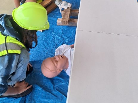 Five young people wearing neon green hard hats and safety vests participate in a hands-on training exercise. Two individuals kneel on a blue tarp, practicing emergency response techniques on a medical manikin, while others observe and take notes around a white folding table.