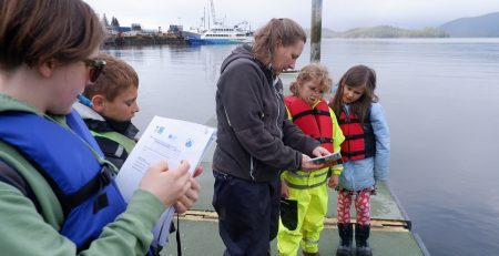 Youths wearing life vests stand on a dock as they fill out forms while an adult explains the process