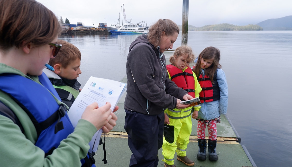 Youths wearing life vests stand on a dock as they fill out forms while an adult explains the process